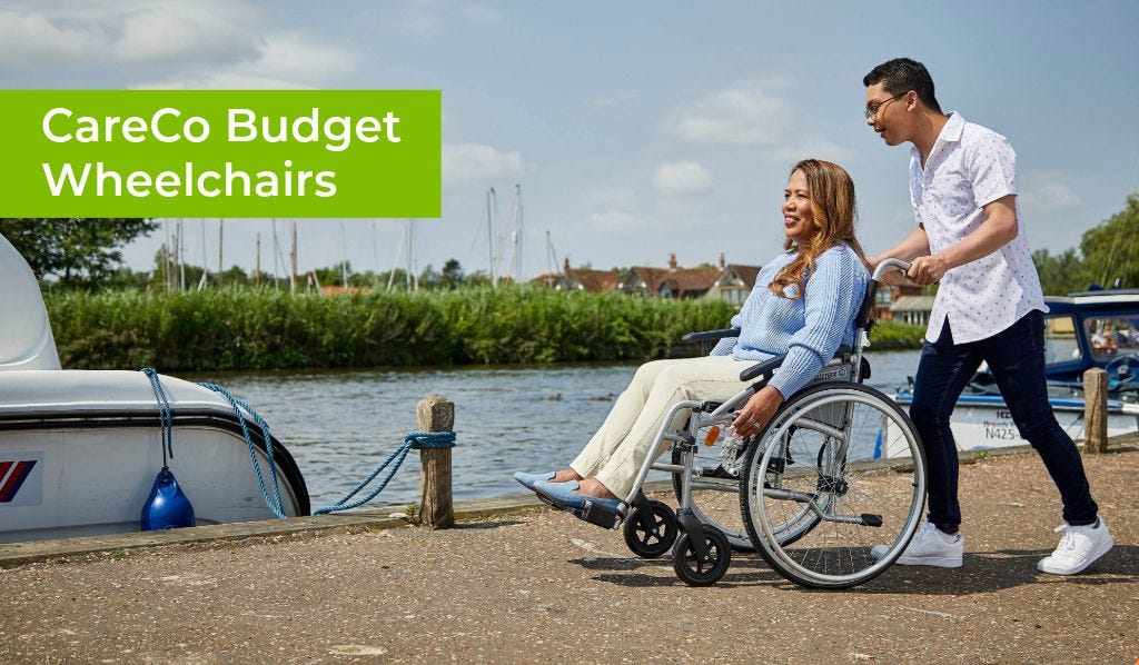Man pushing a woman in a wheelchair by a river with a boat moored