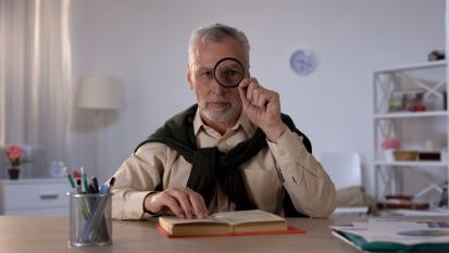 elderly man sat at table with book, looking through magnifying glass