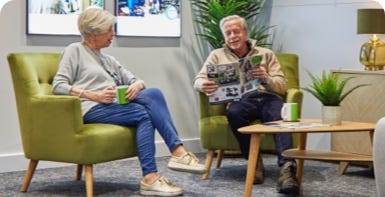 An elderly couple sitting in fireside chairs looking over a brochure together while drinking tea